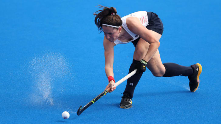 brown-haired woman about to hit a field hockey ball with her stick; water splashes from the field